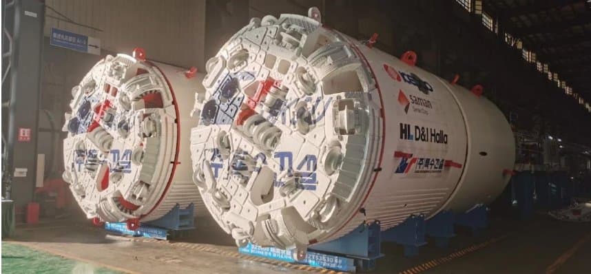Two CRCHI slurry TBM shield bodies lined up side-by-side in the factory hall, painted white with red and blue accents and unit identifiers stencilled on the sides